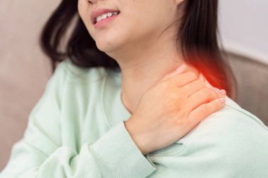 Yeşil kazaklı bir kadın omzunu tutarak acı ve rahatsızlığı, sağlık hizmetlerini ve tıbbi bakım ihtiyacını sembolize eder..