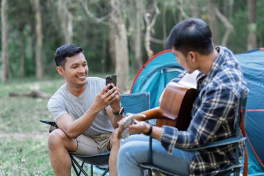 Ormanda çadırlarının yanında kamp yapan neşeli bir çift. Biri gitar çalarken diğeri akıllı telefondan anı yakalıyor..