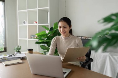 Modern bir ofis ortamında bitki ve minimalist dekorlarla dolu bir dizüstü bilgisayar ve pano kullanan bir iş kadını..