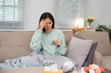 Evdeki kanepede oturan bir kadın, rahat bir battaniyeye sarılmış, elindeki ilaç paketiyle rahatsızlığını giderirken bunalmış görünüyor..