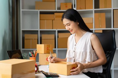 Küçük bir işletme sahibi, kutularla ve ofis malzemeleriyle dolu çalışma alanında çevrimiçi satış siparişleri hazırlayan bir paketin üzerine özenle bir şeyler yazıyor..