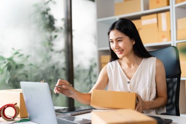 Çevrimiçi siparişleri yönetirken neşeli küçük bir işletme sahibi gülümsüyor, bir paket tutuyor ve masasında kutularla dolu, organize edilmiş bir çalışma alanında çalışıyor..