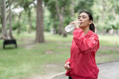 Kırmızı ceketli genç bir kadın yemyeşil bir parkta koşarken su içiyor. Sıhhatinin bir parçası olarak hidrasyon ve zindeliği teşvik ediyor..