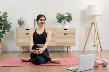 Evde yoga yapan genç bir kadın, dizüstü bilgisayarındaki bir oturumun ardından gülümseyen, spor ekipmanlarıyla çevrili, sağlığını ve farkındalığını destekleyen.
