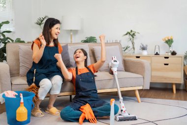 İki kadın temizlik başarılarını sevinçle kutluyor. Etrafı malzemelerle ve vakumla çevrili. Takım çalışmasını ve mutluluğu ön plana çıkarıyorlar..