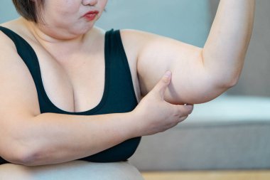 Siyah spor sutyenli bir kadın kolunu inceliyor, vücut bilincini, kendini kabulünü ve sağlık yolculuğundaki sağlığının önemini vurguluyor..