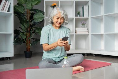 An elderly woman practices wellness by engaging with her smartphone during a yoga session at home, promoting healthy living and mindfulness. clipart