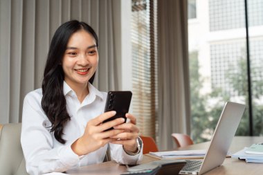 Modern bir ofis masasında, etrafını dokümanlar ve dizüstü bilgisayarlarla çevrili akıllı telefonuyla meşgul olan mutlu profesyonel bir kadın, birden fazla iş becerilerini ve pozitif çalışma ortamını ön plana çıkarıyor..