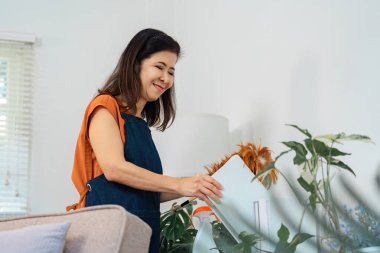 Turuncu gömlekli bir kadın, kitap düzenlerken ve raflarda çiçek düzenlerken gülümsüyor. Etkili ev işleri ve temizlik becerileri sergiliyor..