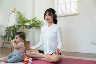Sakin bir anne, kulaklıklarla huzurlu bir ortamda meditasyon yapar. Meraklı bebeğine göz kulak olurken dikkatini korur..