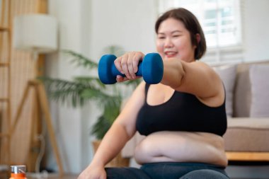 Neşeli, iri yarı bir kadın, evde bir paspasın üzerinde otururken, kendine güvenerek mavi bir dambıl kaldırıyor, spor yolculuğunu ve sağlığını kucaklıyor..