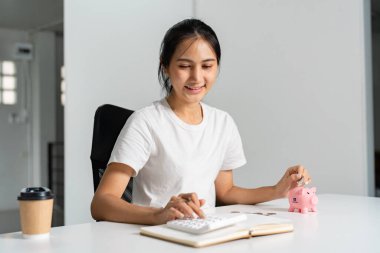 Bütçesini hesap makinesi ve pembe kumbarasıyla yöneten mutlu bir kadın. Çağdaş bir masada otururken kahvesini yudumluyor..