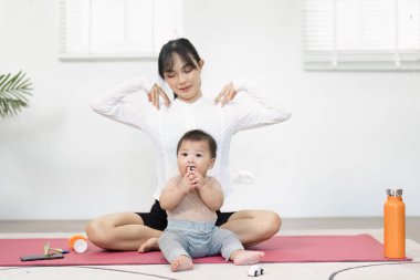 Bir anne yoga minderinin üzerinde nazikçe egzersiz yapar bebeği yanında oturur, oyuncakla mutlu bir şekilde oynar, neşeli ve aktif bir aile hayatı sergiler..
