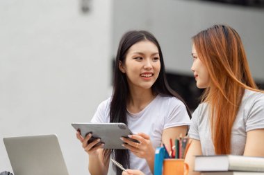 İki öğrenci fikirlerini paylaşıyor ve bir tablet üzerinde işbirliği yapıyor, akademik uğraşlarında takım çalışması ve yaratıcılık sergiliyor, modern eğitim araçlarını vurguluyor.