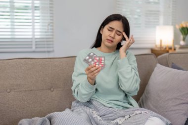 Bir kadın, battaniyeye sarılı bir şekilde elinde ilaç tutarken rahatsızlık hissediyor. Rahatsızlık anlarında, sıcacık bir evde kendine bakmanın ve sağlığın önemini vurguluyor..