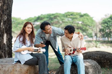 Üç arkadaş parkta keyifli bir anı paylaşıyor, ukulele oynayıp birlikte gülüyorlar, canlı bir arkadaşlık ve yaratıcılık ortamı yaratıyorlar..