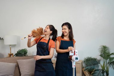 Önlüklü iki kadın arasında mutlu bir etkileşim var. Biri elinde toz bezi tutarken diğeri de elektrikli süpürgeyle salonda duruyor..