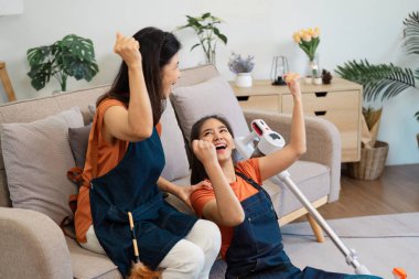 Excited expressions as two women share a moment of victory over their cleaning tasks, sitting on a couch with a vacuum cleaner and a feather duster nearby. clipart