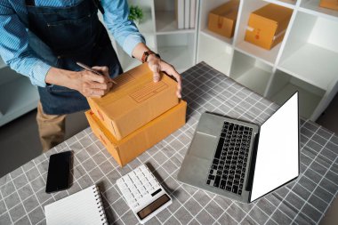 Yanında dizüstü bilgisayarı ve hesap makinesiyle paketleri etiketleyen yaşlı bir adam modern çalışma alanındaki titiz organizasyon ve lojistik işlemlerini gösteriyor..