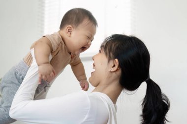 Bir anne mutlu bir şekilde bebeğini kucağına alır, her ikisi de evde neşeli bir anda kahkahalarını paylaşır, aile bağının ve sevginin neşesini gösterir..