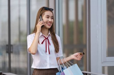Gülümseyen ve telefonuyla konuşan mutlu genç bir kadın, alışveriş torbaları ve alışveriş merkezinde bir kredi kartı tutuyor..