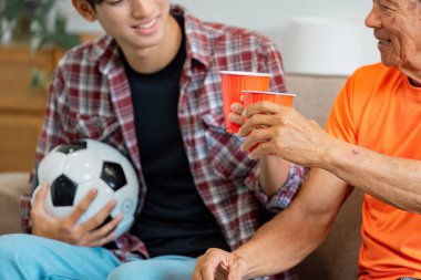 Bir futbol topu tutarken kırmızı bardaklarla kadeh kaldıran genç bir adam ve dedesinin yakın çekimi, maç sırasında neşeli bağlarını gösteriyorlar..