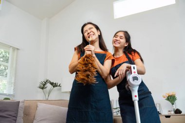 İki turuncu tişörtlü kadın temizlik yaparken eğleniyor, biri toz bezi tutuyor, diğeri ise süpürge, ev işleri ve ev idaresinde takım çalışması yapıyor..