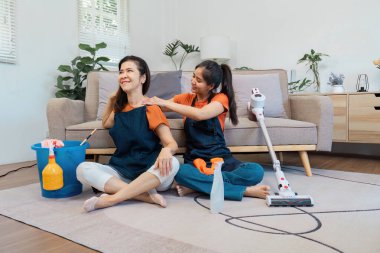 Turuncu gömlekli iki kadın yerde oturuyor, biri temizlik malzemeleri ve ev işlerinde arkadaşlığı ön plana çıkarırken diğerinin önlüğünü şakaya vuruyor..