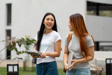 İki farklı üniversite öğrencisi kampüste birlikte yürüyor, kitap ve dizüstü bilgisayar taşırken gülüyor ve eğleniyorlar..