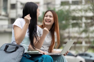 İki farklı üniversite öğrencisi dışarıda başarılı bir çalışma seansını kutluyorlar, yanlarında bir dizüstü bilgisayarla sevinç ve coşku ifade ediyorlar..