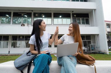 İki farklı kız öğrenci başarılı bir anı kutlarken kampüste bir bankta oturup neşe ve arkadaşlığı paylaşıyorlar..