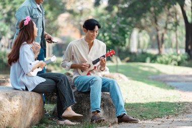 Bir kayanın üzerinde oturan, ukuleles çalan ve güzel bir açık hava parkında birlikte müziğin keyfini çıkaran bir grup arkadaş..