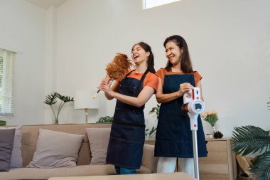 Önlüklü iki kadın, oturma odalarını temizlerken tüy fırçası ve elektrikli süpürgeyle gülüp poz veriyorlar..