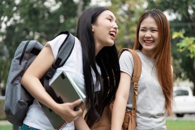 Kampüste birlikte yürüyen iki farklı üniversite öğrencisi sırt çantaları ve okul malzemeleri taşırken birbirlerine gülüp eğleniyorlar..