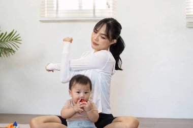 Asyalı bir anne, bebekle uğraşırken evde esneyerek, spor ve aktif ebeveynlik dengesini göstererek, refahı kucaklar..
