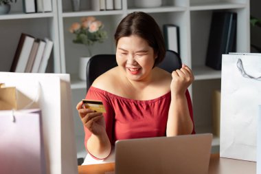 A delighted woman celebrates her online shopping success, holding a credit card while surrounded by shopping bags. Ideal for themes of e-commerce and retail joy. clipart