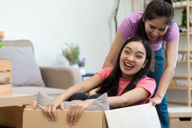 İki kadın taşınırken keyifli bir anı paylaşır, biri bir kutuda oturur ve diğeri onu şakayla iter, taşınma ve arkadaşlığın heyecanını vurgular..