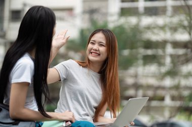 İki öğrenci gülümsüyor ve bir dizüstü bilgisayarla otururken beşlik çakıyor, açık hava eğitim ortamında neşeli bir takım çalışması ve yoldaşlık anı sergiliyorlar..