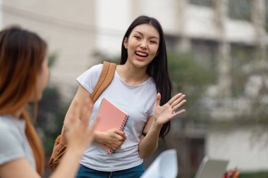 Çeşitli bir üniversite öğrencisi, elinde bir defter tutarken arkadaşına gülümseyip el sallıyor ve dışarıda ders çalışmaktan zevk alıyor..