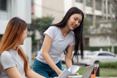 İki farklı üniversite öğrencisi açık havada etkin bir şekilde bir çalışma seansına katılıyor, belgeleri paylaşıyor ve bir dizüstü bilgisayarın yanında işbirliği yapıyorlar..