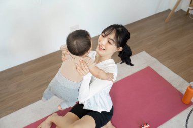 Bir anne neşeli bir şekilde bebeğini havaya kaldırır, kapalı bir alanda yoga minderinde neşeli ve sevgi dolu bir etkileşimi paylaşır..