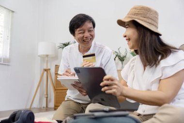 An Asian couple shares smiles while discussing their online shopping plans, with one partner holding a credit card and the other using a tablet for purchases. clipart