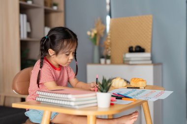 Küçük bir masada oturan kararlı genç bir kız, modern bir yaşam alanında notlar ve sanatsal malzemelerle çevrili, özenle harflerini ve çizimlerini çalışıyor..
