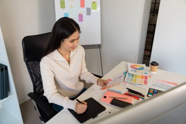 Yaratıcı bir kadın tasarımcı tabletine renk fikirleri çiziyor ve beyin fırtınası yapıyor. Modern ofis düzeninde çeşitli renk örnekleriyle çevrili..