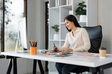 Neşeli bir bayan tasarımcı, şık modern ofis ortamında renk paletleri ve kırtasiye malzemeleriyle çevrili tableti üzerinde çalışırken gülümsüyor..