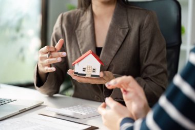 Bir emlakçı müşterisine model ev tanıtıyor, anahtar özellikleri anlatıyor ve profesyonel ofis ortamında finansman seçeneklerini tartışıyor..