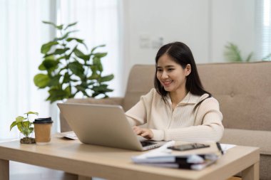 Neşeli genç Asyalı bir kadın masasında oturuyor, mutlu bir şekilde dizüstü bilgisayarında çalışıyor, etrafı parlak ve üretken bir ev ortamıyla çevrili..