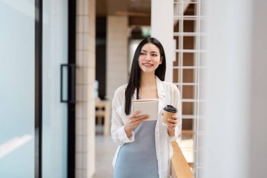 Tabletli ve kahveli genç bir kadın modern profesyonelliği ve çoklu görevi temsil eden çağdaş bir ofiste yürürken gülümsüyor..
