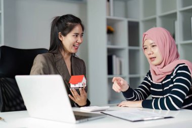 Bir emlakçı ve bir müşteri, şık bir ofis ortamında model bir evi referans olarak kullanarak mülkiyet seçenekleri hakkında bir tartışmaya girişirler..