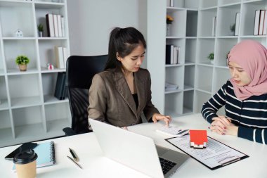 Profesyonel bir emlakçı, lüks bir ofis ortamındaki bir müşteriyle istişare sırasında mülk maliyetlerini değerlendirmek için hesap makinesi kullanır..